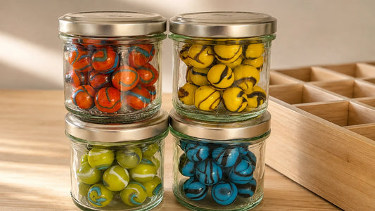 Quatre bocaux en verre à couvercle métallique, empilés deux par deux, chacun rempli de billes MesBilles d'une seule couleur (orange, jaune, vert, bleu), sur un plan de travail en bois éclairé en lumière naturelle, à côté d'un casier à compartiments