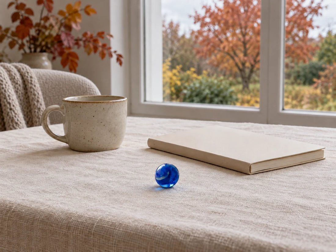 Une bille de verre bleue posée sur une nappe de lin écru, près d'un cahier fermé et d'une tasse en grès, devant une fenêtre ouverte sur un jardin aux arbres roux.