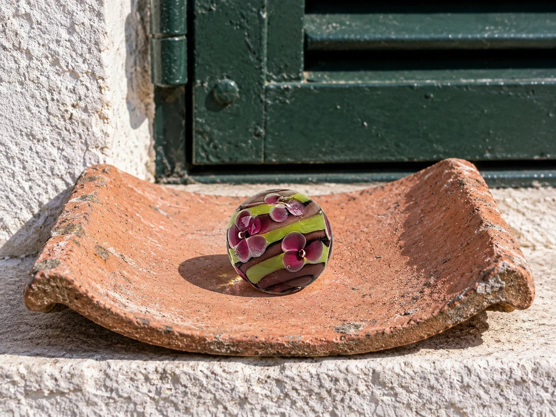 Une bille de verre rayée de brun et de vert anis, ornée de fleurs roses, posée dans une tuile canal en terre cuite sur le rebord d'une fenêtre, devant un volet vert foncé.